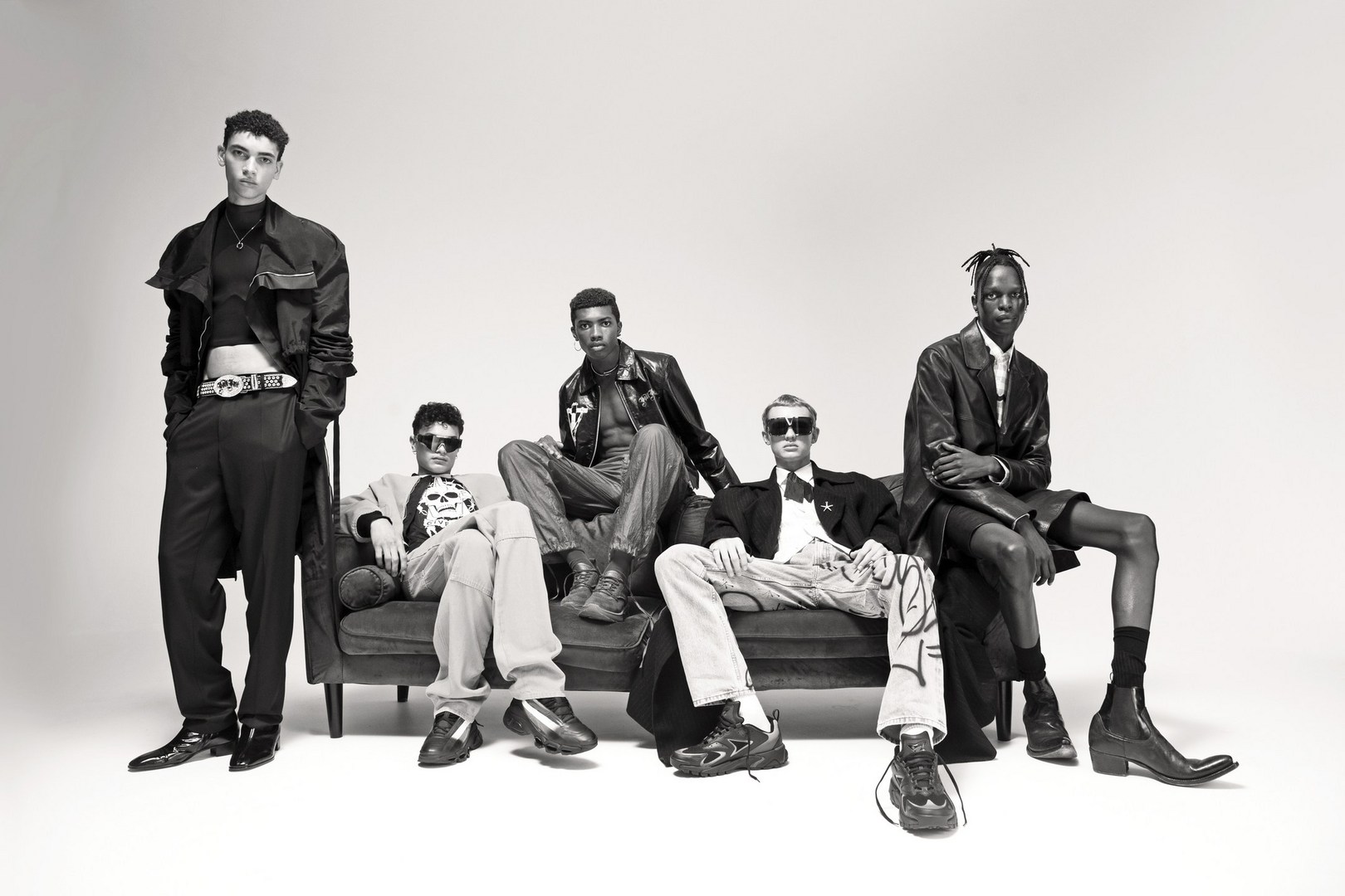 Five fashionably dressed young adults of varying ethnicities pose on a dark sofa in a studio setting with a light background.