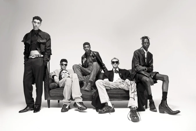 Five fashionably dressed young adults of varying ethnicities pose on a dark sofa in a studio setting with a light background.