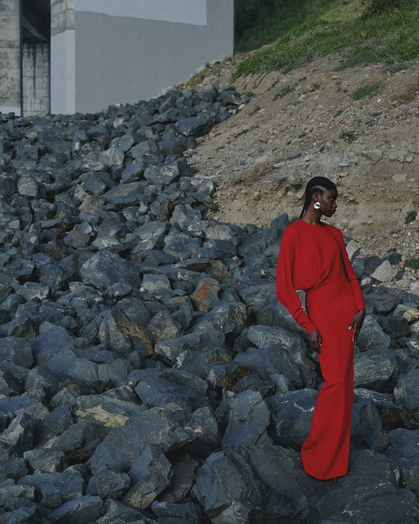 Woman in a long red dress poses on a rocky landscape with a neutral concrete structure in the background, fashion, beauty, outdoor shot.