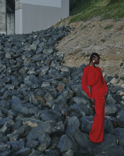 Woman in a long red dress poses on a rocky landscape with a neutral concrete structure in the background, fashion, beauty, outdoor shot.