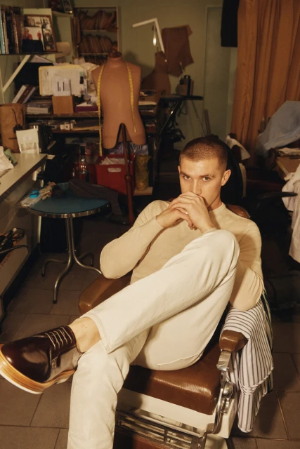 Man in cream sweater and light jeans sits in a barber chair in cluttered room, looking at the camera with hands together.