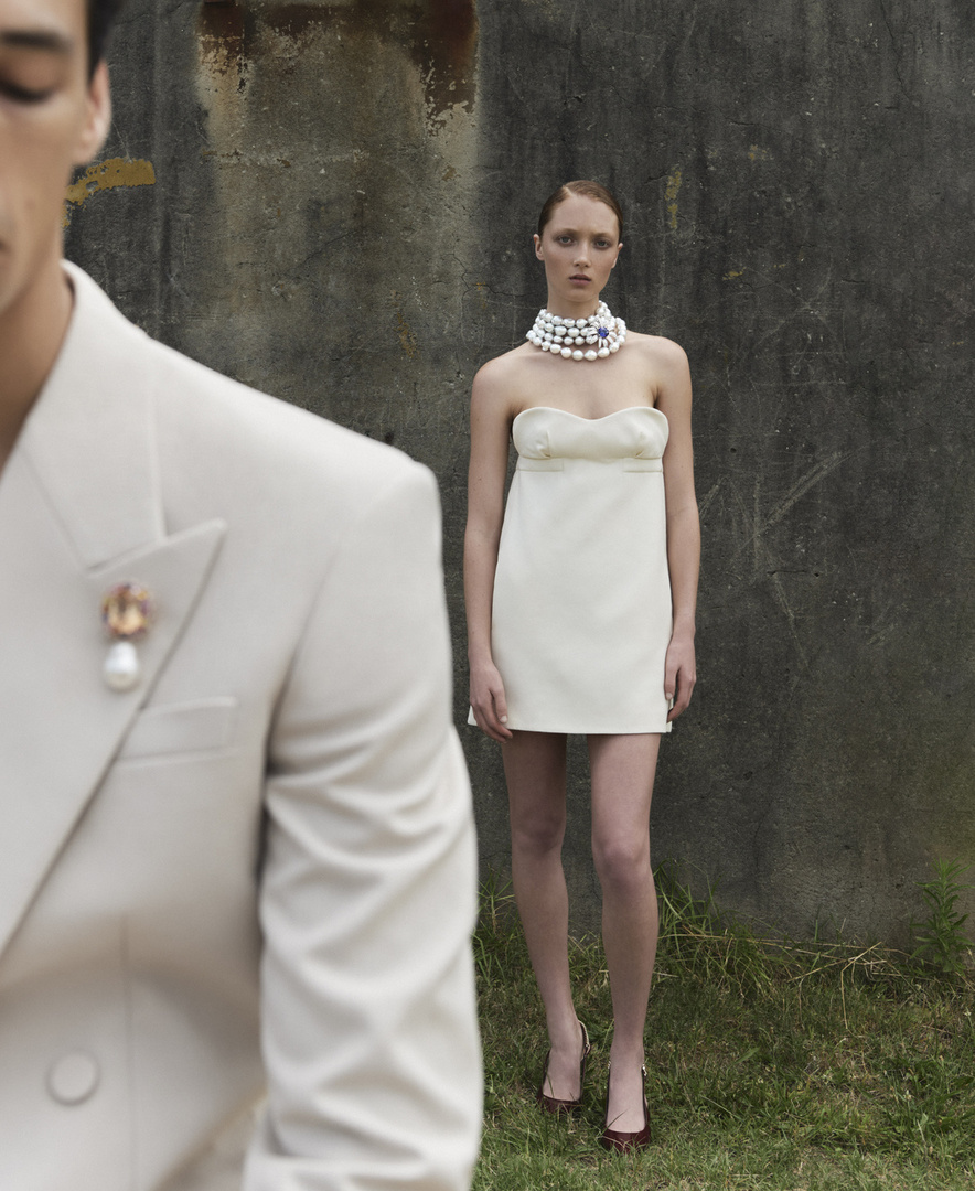 A young woman wearing a cream-colored mini dress and pearl necklace stands outdoors near a man in a cream suit and pearl brooch.