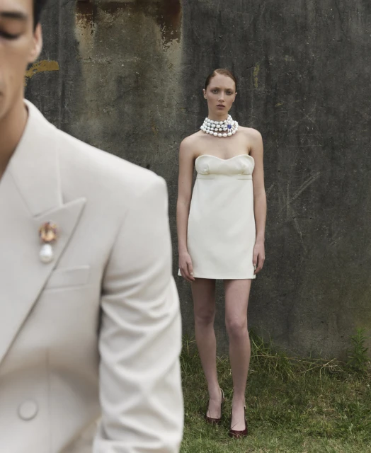 A young woman wearing a cream-colored mini dress and pearl necklace stands outdoors near a man in a cream suit and pearl brooch.