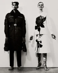 Monochrome fashion editorial featuring a male model in a dark coat and a female model in a white floral outfit against a plain background.