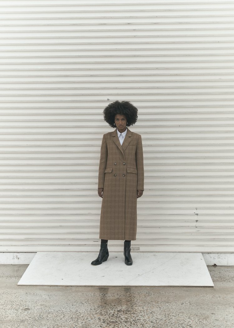 A single adult female model with afro hairstyle poses in an outdoor setting, wearing a tan plaid overcoat and black ankle boots against a white wall.