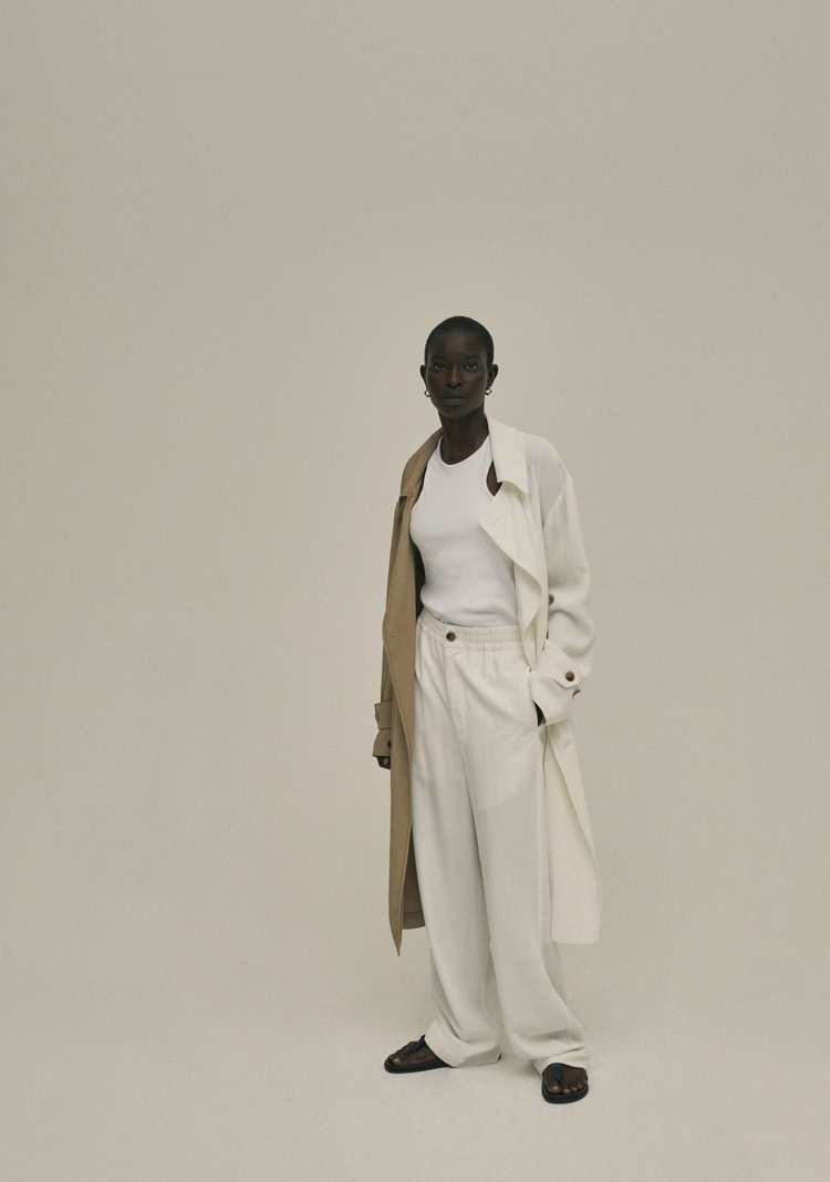 A fashion photo of an adult female model in a white top, white pants, and a trench coat, standing in front of a neutral background.