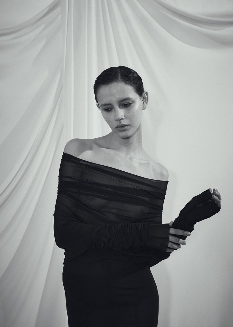 Monochrome portrait of a young woman in a black off-the-shoulder dress posing in front of a draped white fabric backdrop.
