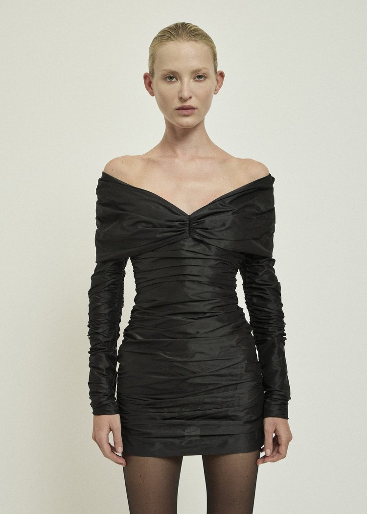 A fair-skinned woman posing in a short, ruched, off-the-shoulder black dress in a studio setting, lit by even, diffused lighting.