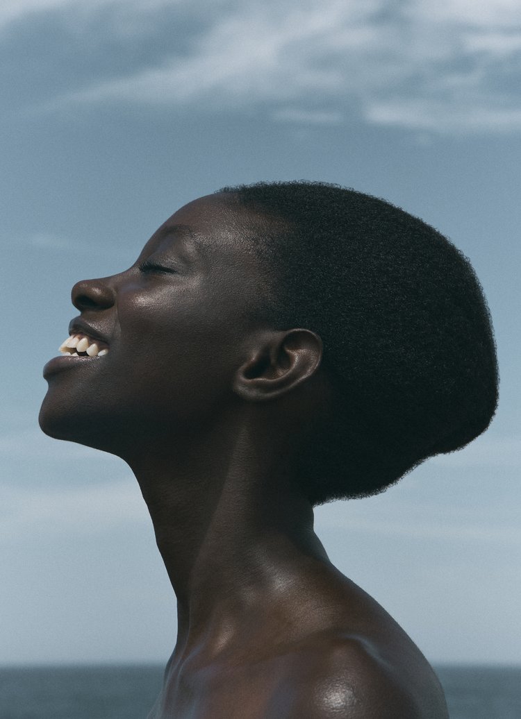 Close-up portrait of a smiling African woman with dark skin, looking up against a blue sky with clouds and a glimpse of dark water.