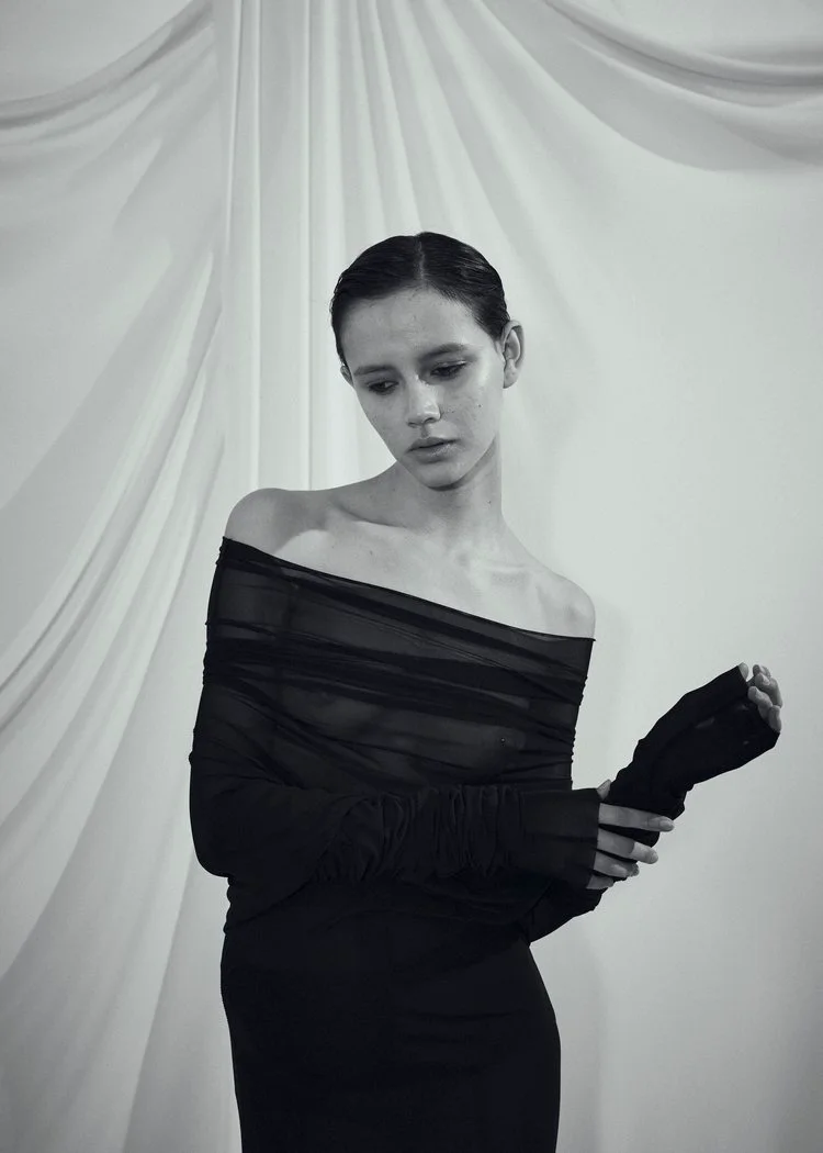 Model in a black off-the-shoulder draped top posing against a softly folded white fabric backdrop in a black and white portrait.