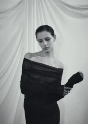 Model in a black off-the-shoulder draped top posing against a softly folded white fabric backdrop in a black and white portrait.