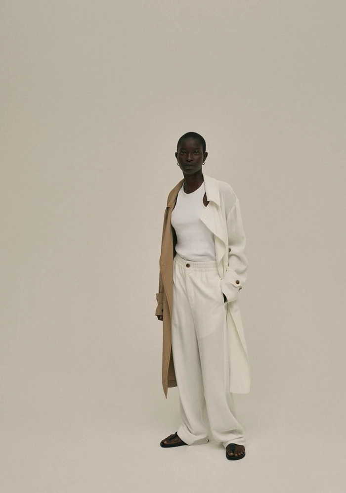 Model wearing a white tank top, cream trousers, and a layered trench coat against a plain neutral background.