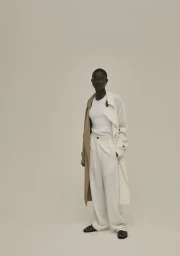 Model wearing a white tank top, cream trousers, and a layered trench coat against a plain neutral background.