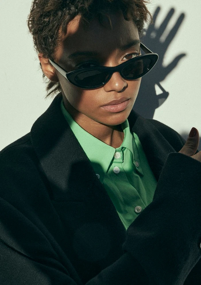 Portrait of a model wearing black cat-eye sunglasses, a vibrant green shirt, and a dark coat against a plain background.