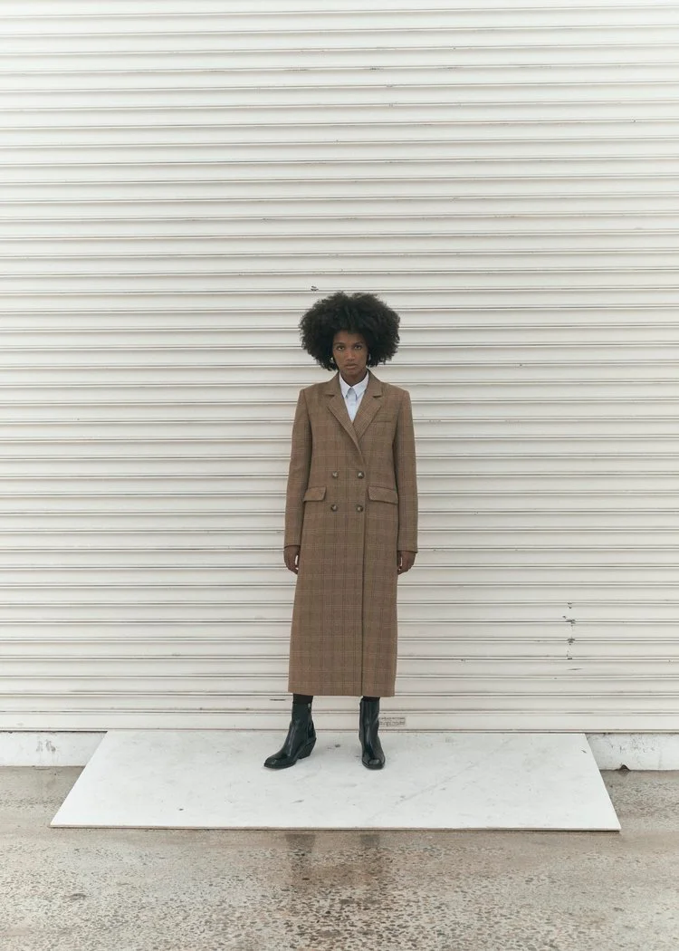 Model standing in front of a white roller shutter door wearing a long brown plaid coat and black boots.