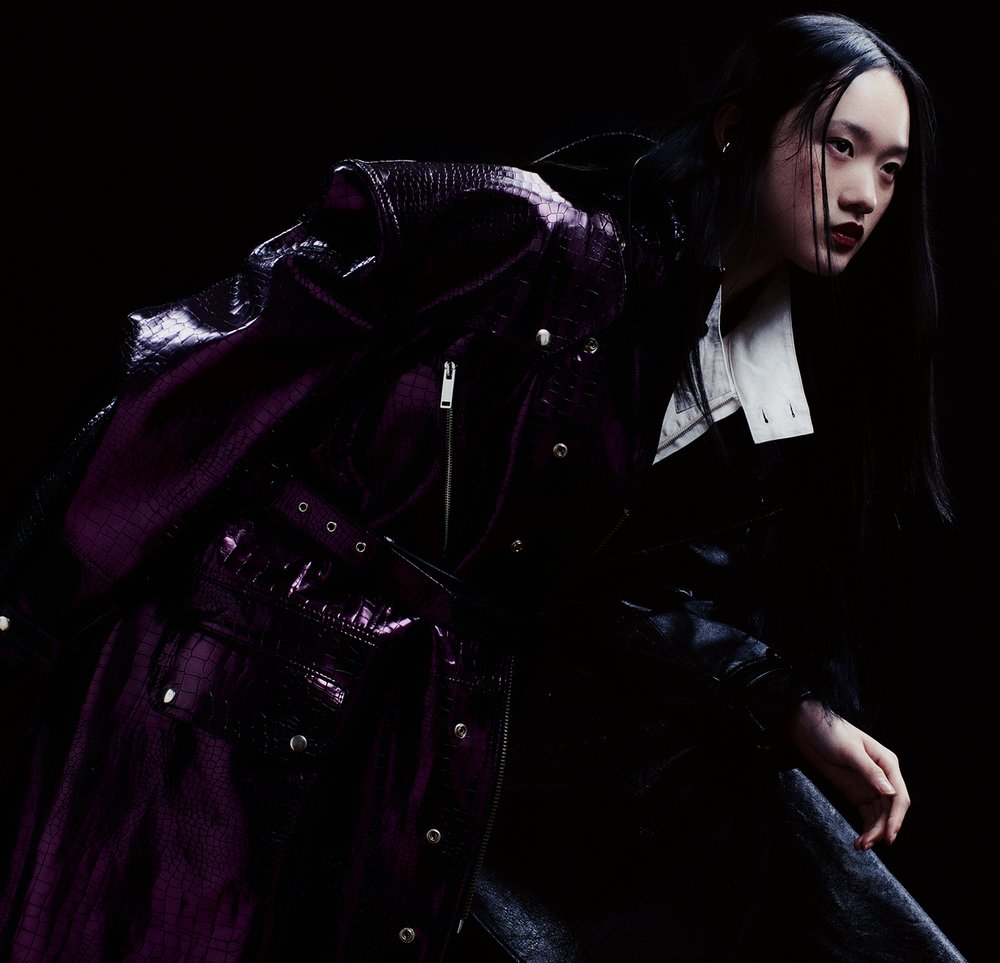 A stylish Asian woman poses in a purple crocodile-pattern coat with a white collared shirt in a dark studio setting.