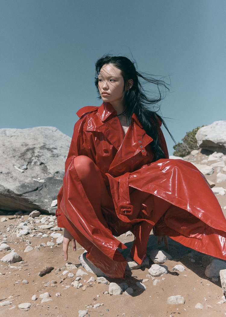Woman in a red patent leather trench coat and pants crouching on a rocky desert landscape under a clear blue sky in a fashion editorial.