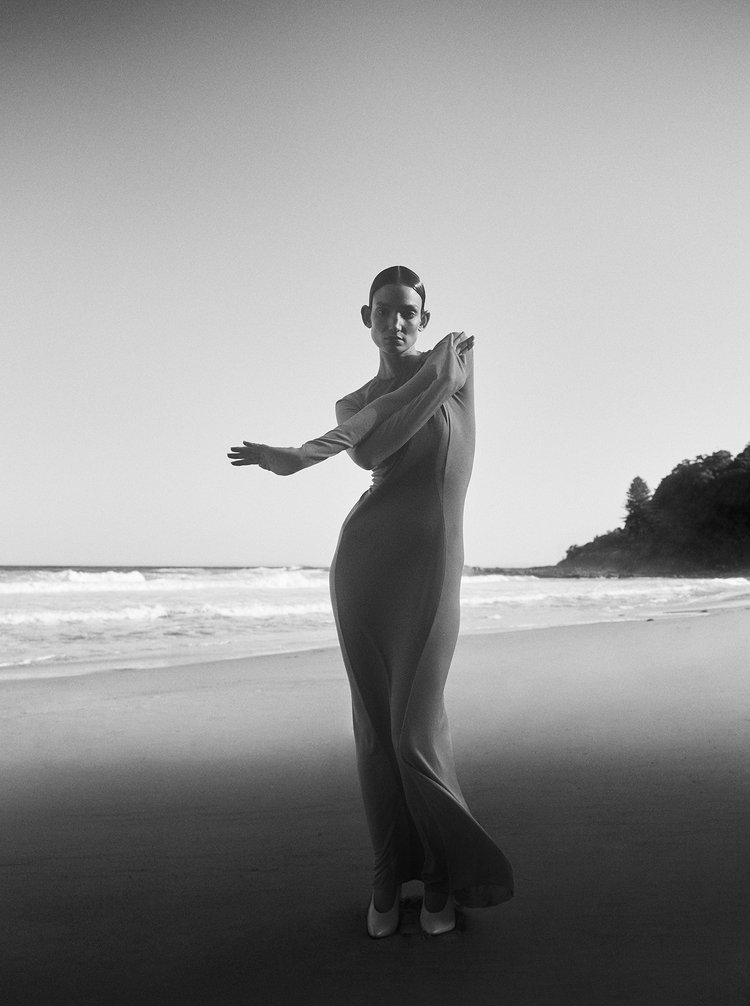 Monochrome image of a slender woman in a long dress posing on a sandy beach with gentle waves and a distant, tree-covered headland.