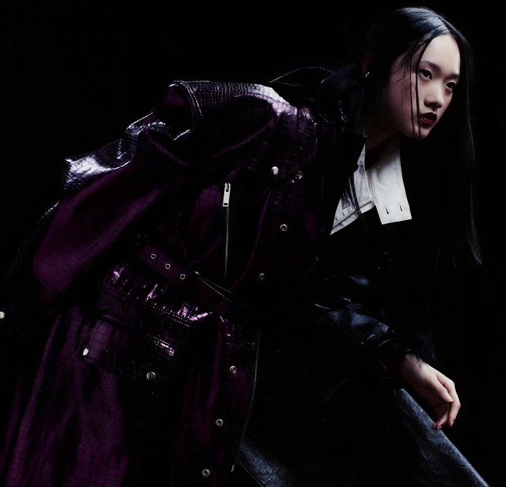 Model in glossy purple textured coat and white shirt against a dark background, styled by Cheryl Tan