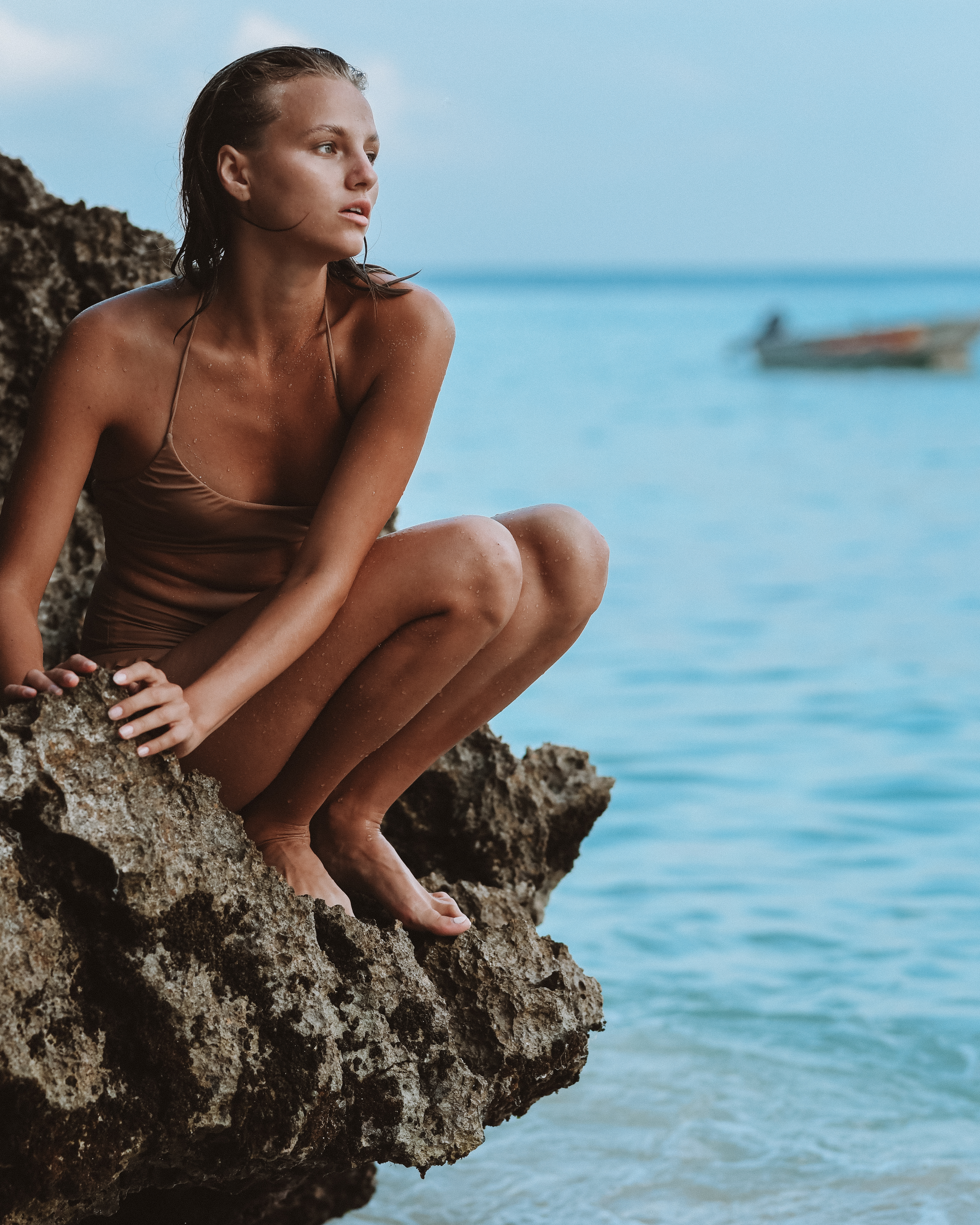 Woman in a swimsuit crouches on a rocky outcrop by the sea. She looks out at the ocean with a boat visible in the distance.