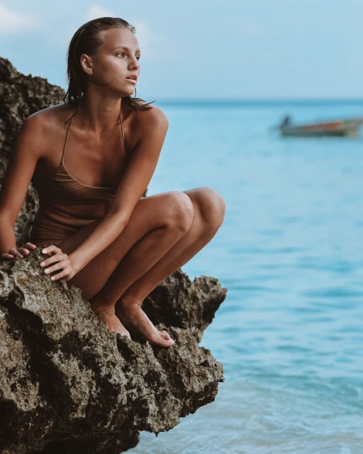 Woman in a swimsuit crouches on a rocky outcrop by the sea. She looks out at the ocean with a boat visible in the distance.