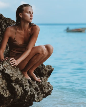 Woman in a swimsuit crouches on a rocky outcrop by the sea. She looks out at the ocean with a boat visible in the distance.