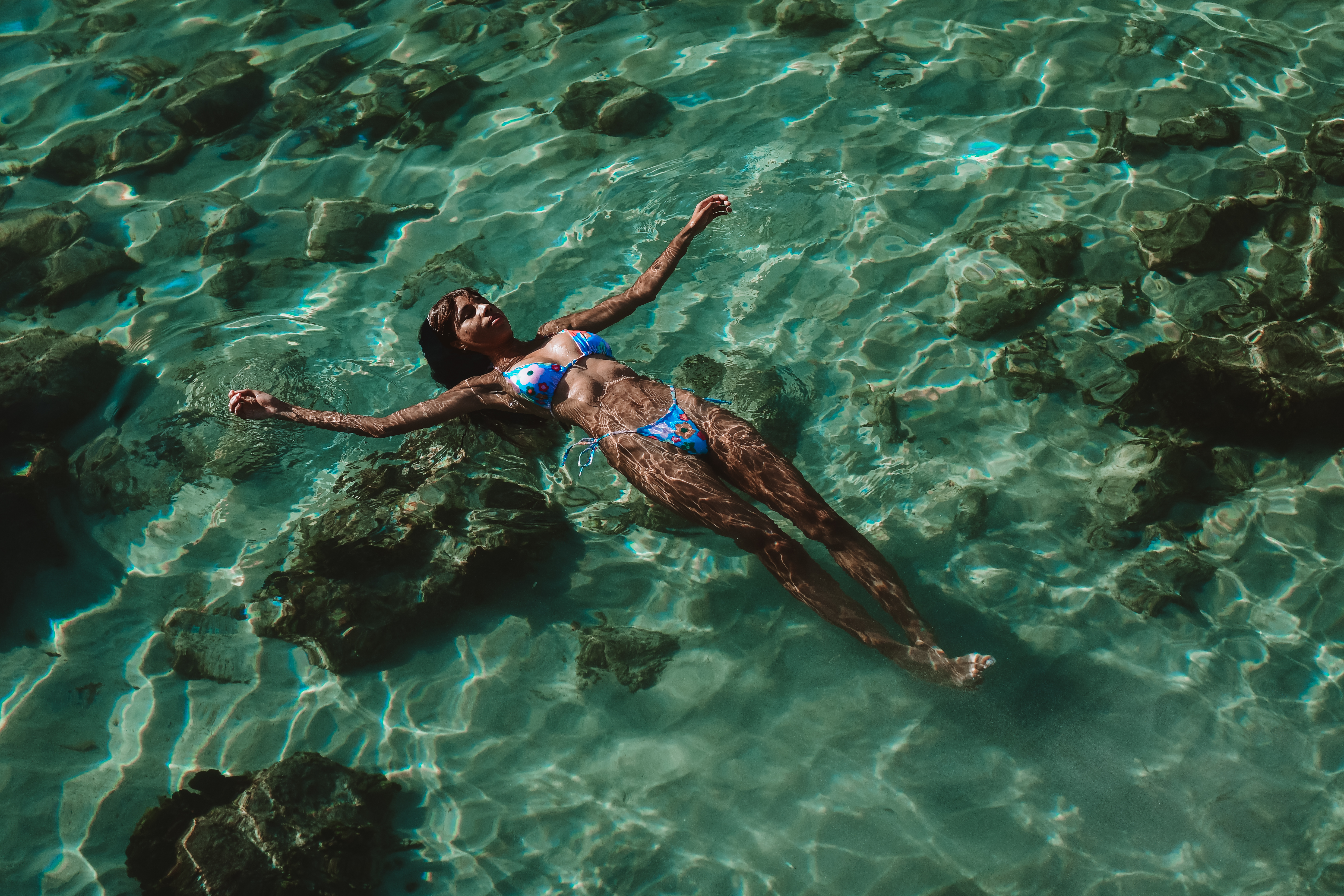 Woman in blue bikini floating in clear, turquoise water with submerged rocks visible underneath, basking in bright sunlight.