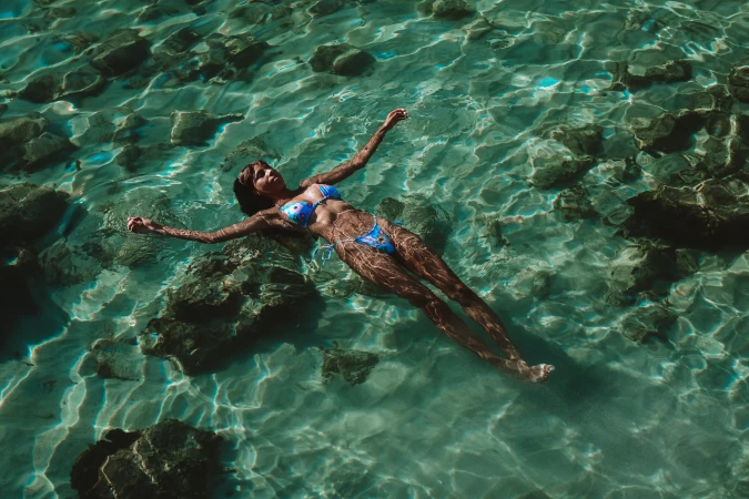 Woman in blue bikini floating in clear, turquoise water with submerged rocks visible underneath, basking in bright sunlight.