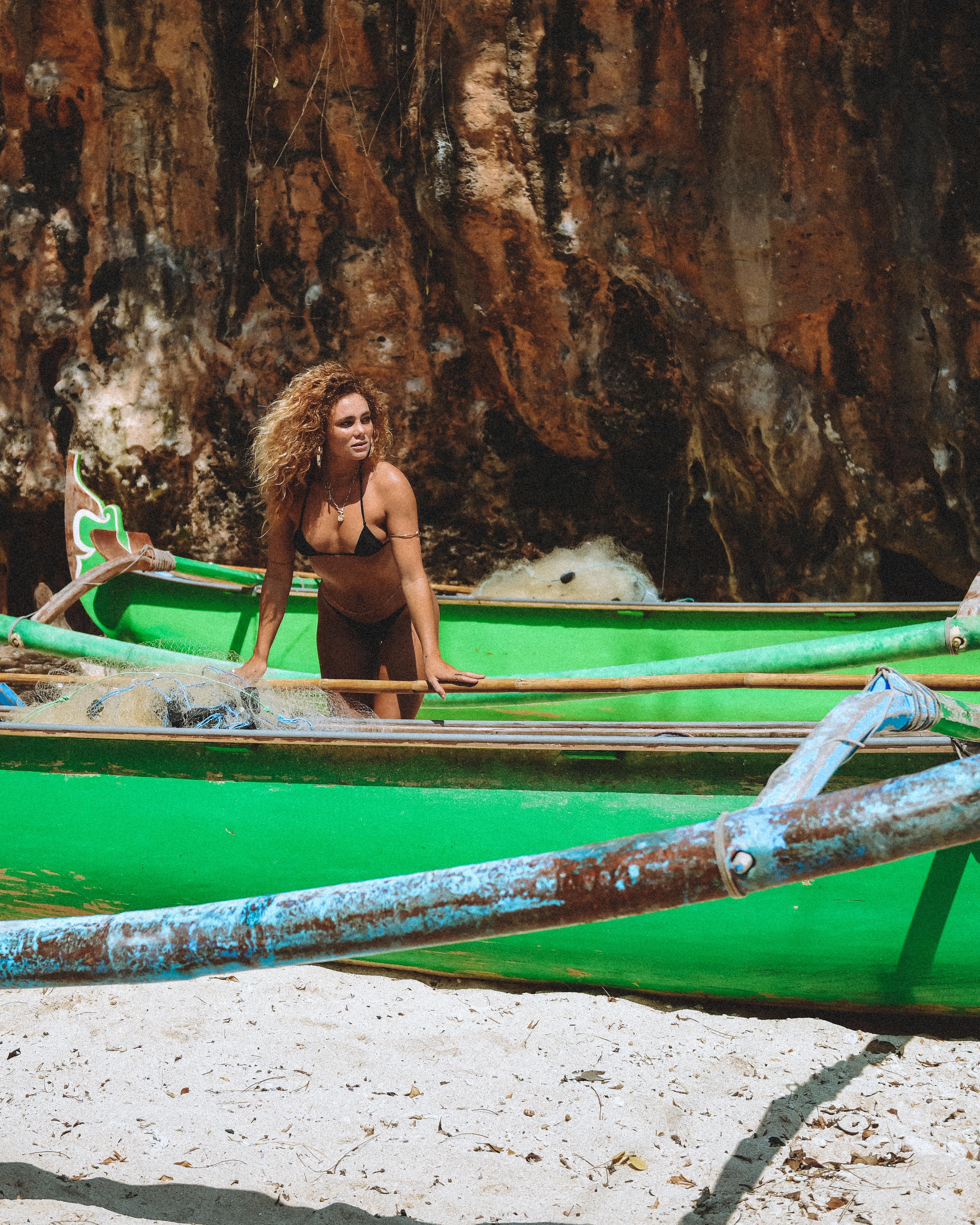 Adult woman in black bikini stands in weathered green rowboat on a sandy beach with a large rocky outcropping in the background.