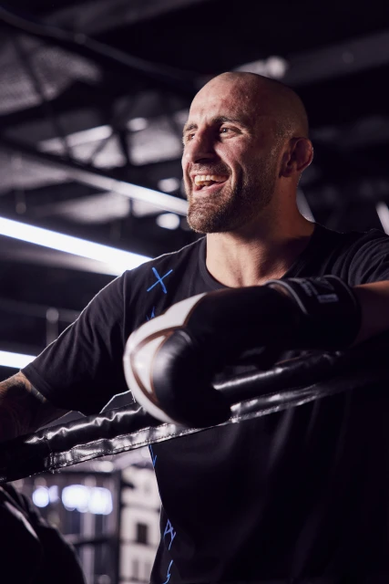 Smiling male boxer in a black t-shirt with boxing gloves, leans against the ropes in a boxing ring. Dark, indoor setting.