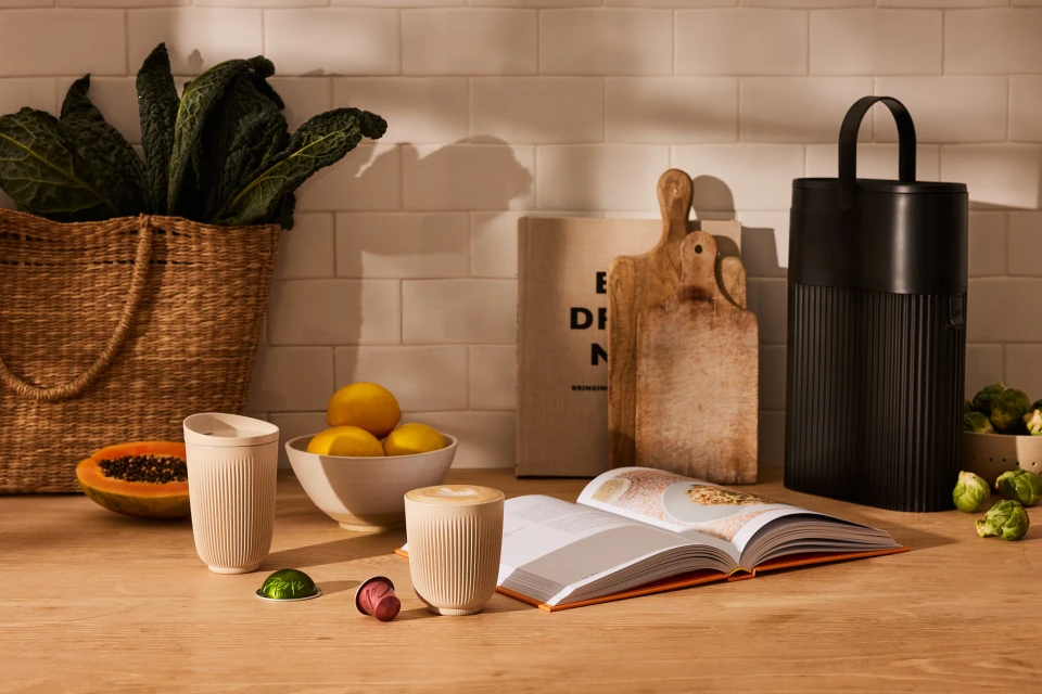 Still life of coffee cups, cookbook, and fresh produce on a wooden countertop against a white brick wall backdrop in a kitchen setting.