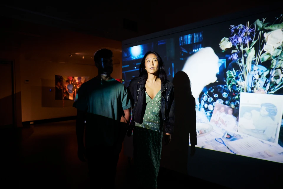 A woman in a dress and jacket stands near a man in an art gallery, illuminated by a projector showing a still life image.