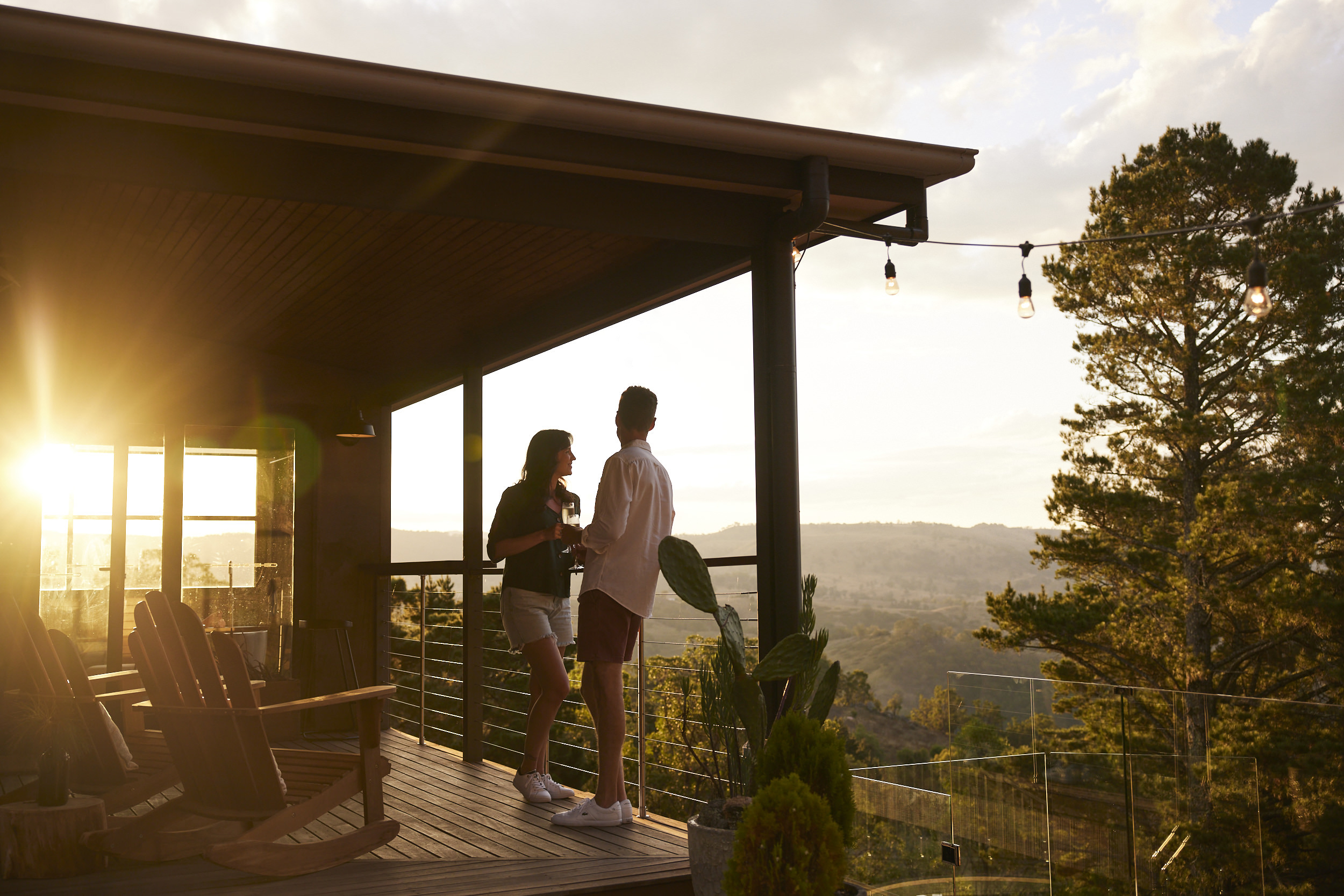 A man and woman standing together with drinks on a timber deck overlooking rolling hills at golden hour sunset.
