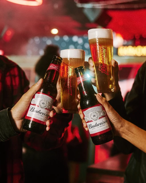 People toasting with Budweiser beer bottles and glasses filled with beer, indoors, red lighting and a blurred background.
