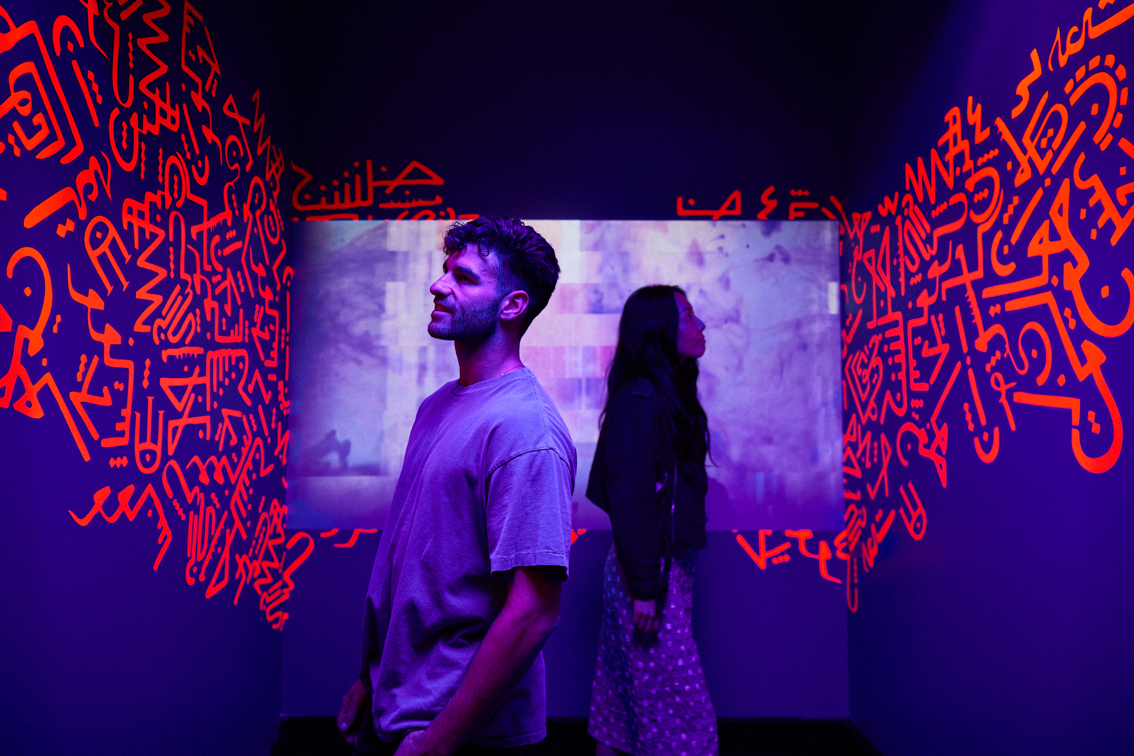 Two people in a dark room with orange Arabic graffiti on the walls, looking at a screen with abstract imagery, bathed in purple light.