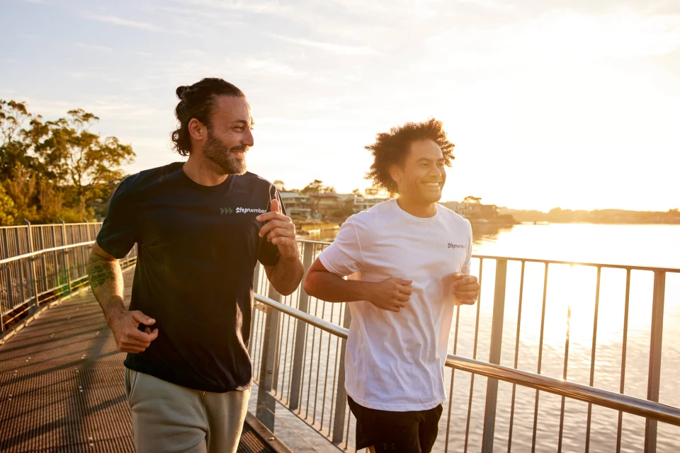 Two athletic adult men jogging on a bridge at sunset near a lake, wearing branded t-shirts and sportswear. Fitness and outdoor lifestyle.