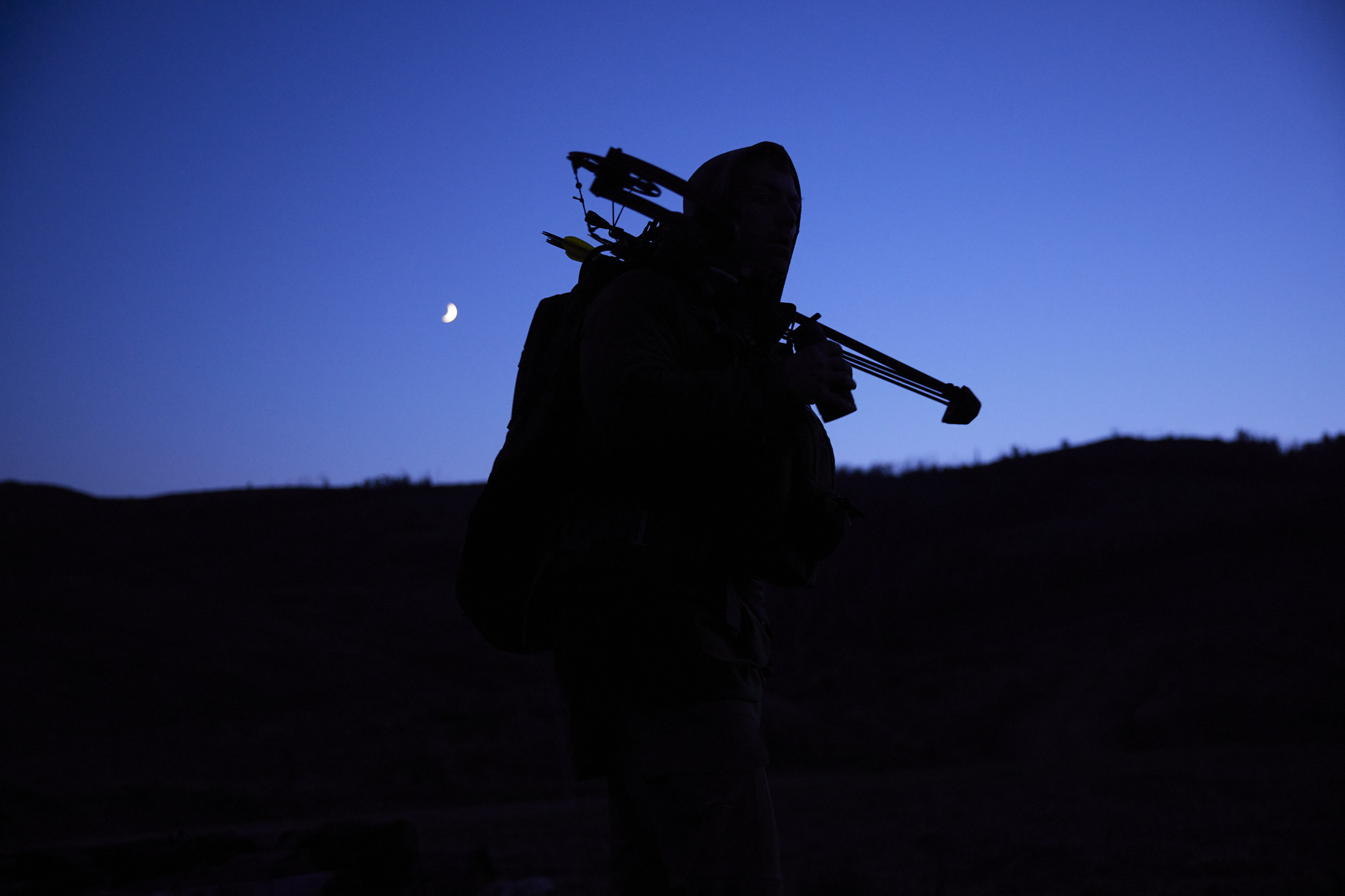 Silhouette of person with crossbow stands against dark landscape with moon in the sky, captured at night with a minimalist composition.