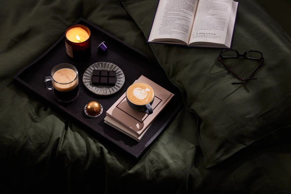 Still life of coffee, chocolate, books, glasses, and candle on a dark wooden tray, set on a dark green bedsheet indoors.