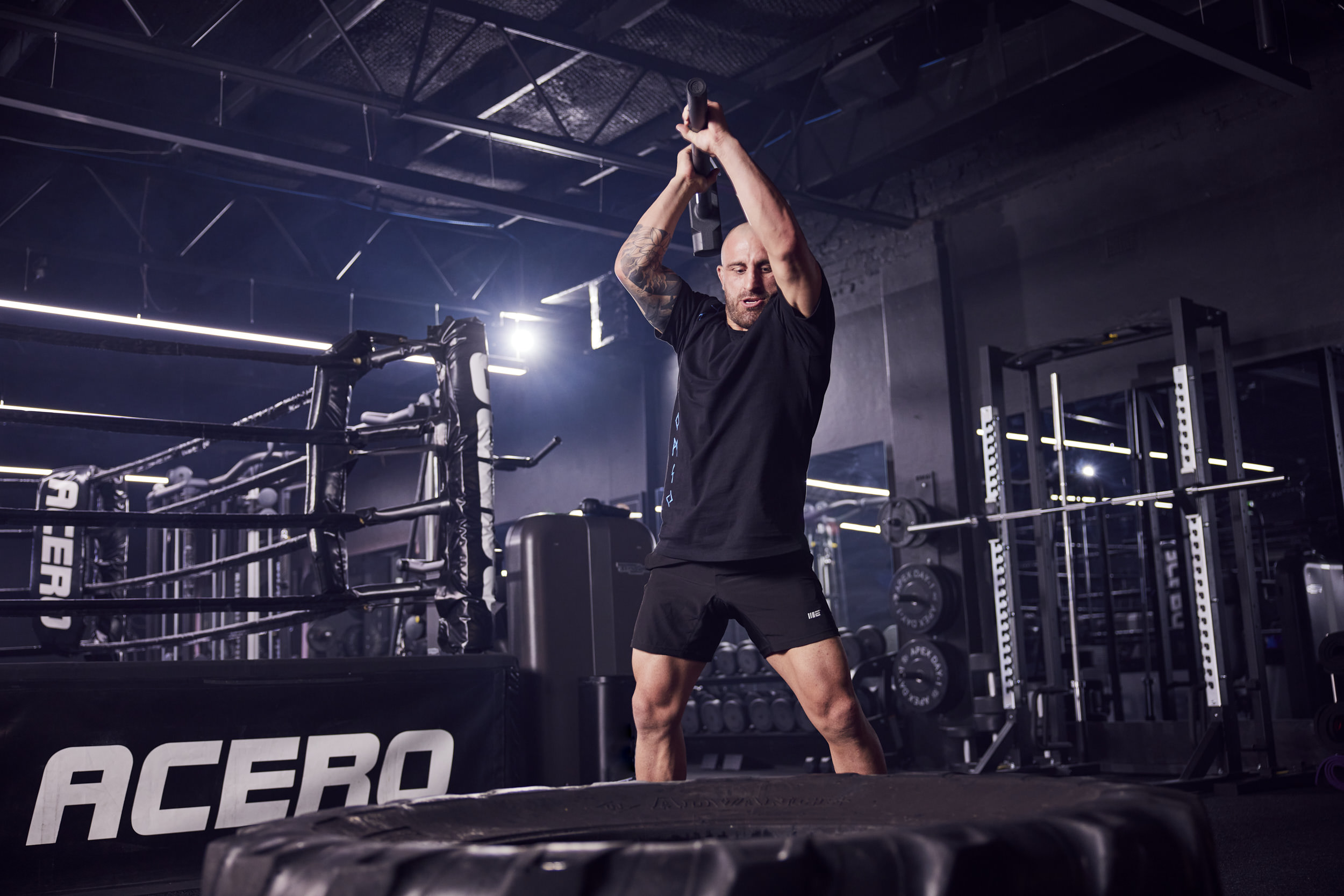 Muscular man in sportswear powerfully swings sledgehammer at tire in dark indoor gym setting with weights and boxing ring equipment.