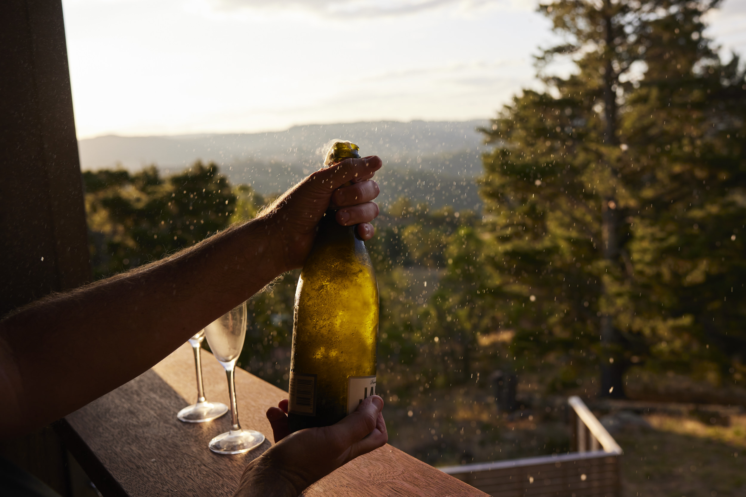 Champagne bottle being opened outdoors on a wooden balcony, with two champagne glasses and forest and mountain views in the background.