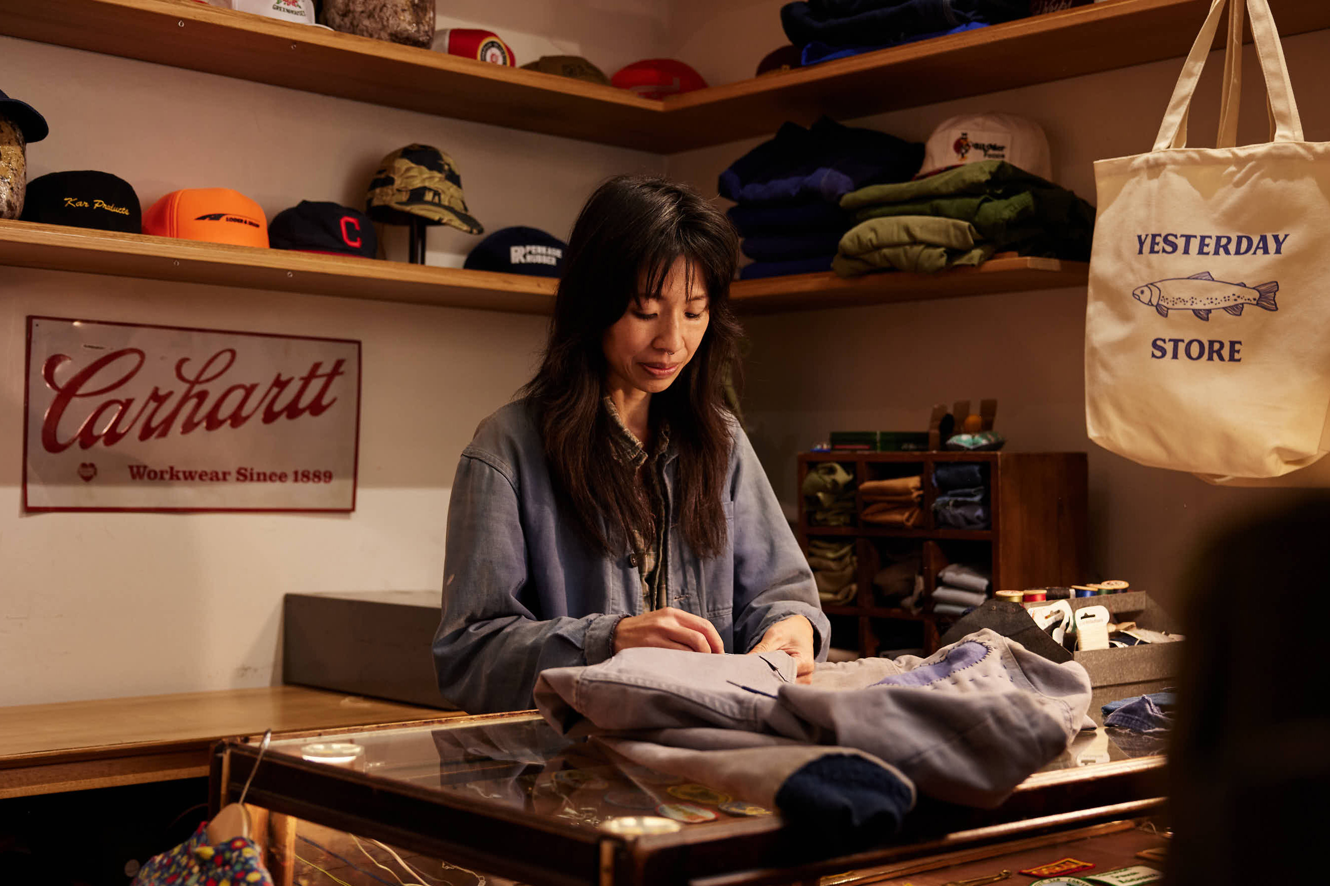 Adult woman tending a counter in a clothing store, surrounded by garments, hats, and accessories in a warm, inviting atmosphere.