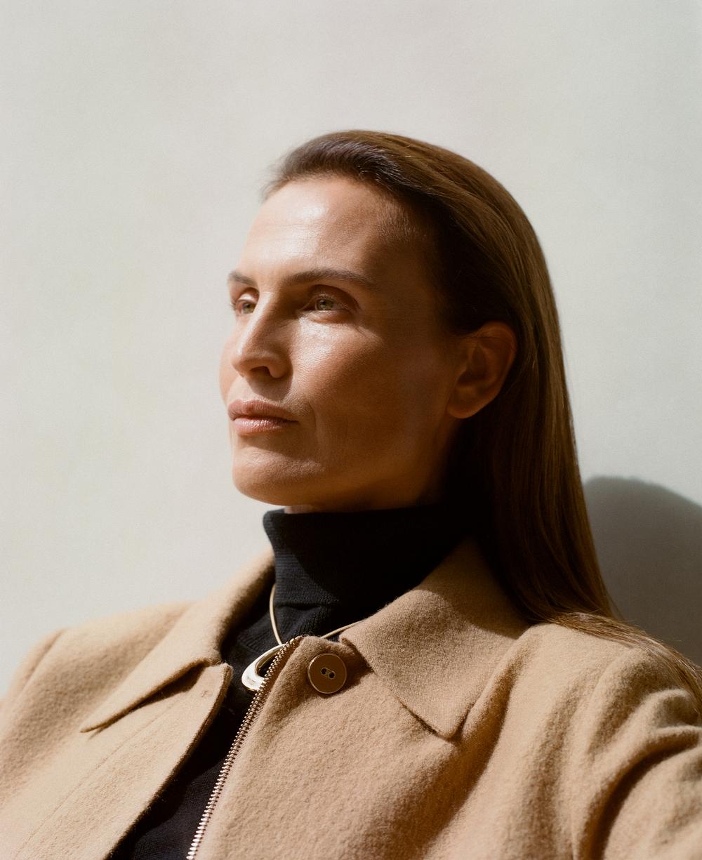 Portrait of a stylish woman with long brown hair, wearing a black turtleneck and tan wool coat, posing against a neutral background.