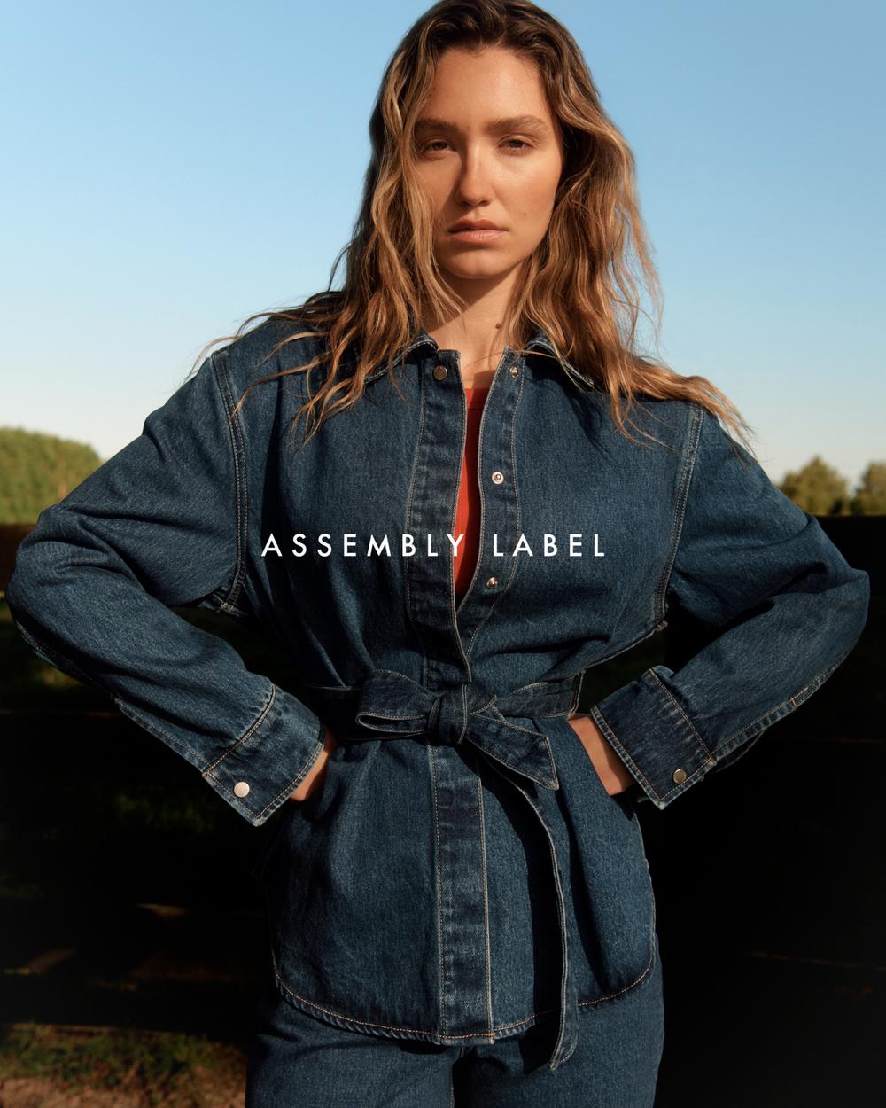 A woman in a denim outfit with hands on hips stands outside, posing in an eye-level medium shot with soft natural lighting.