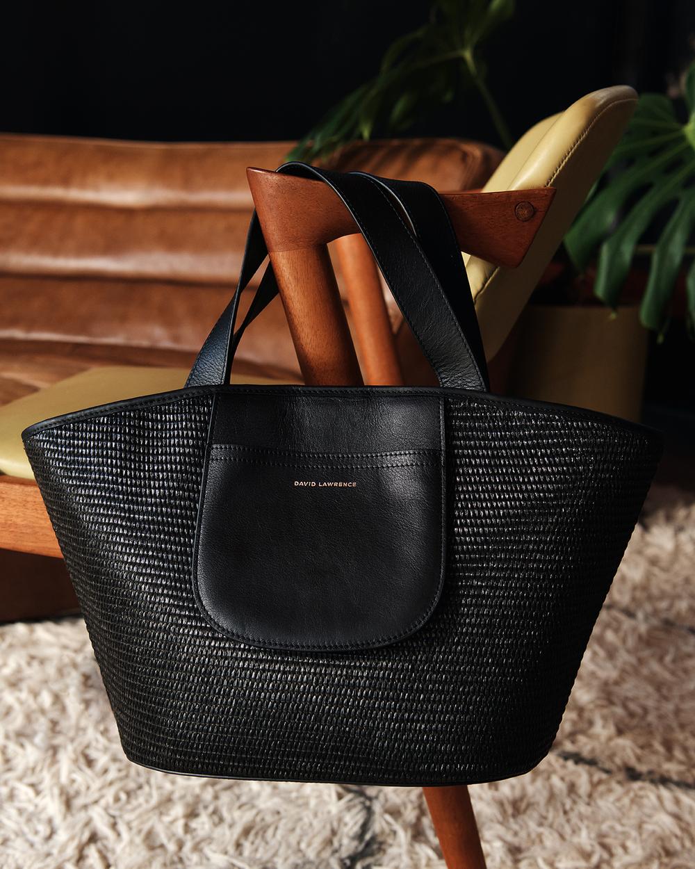 Black woven handbag with leather trim on a tan chair against a dark background with a potted plant, providing a stylish product shot.