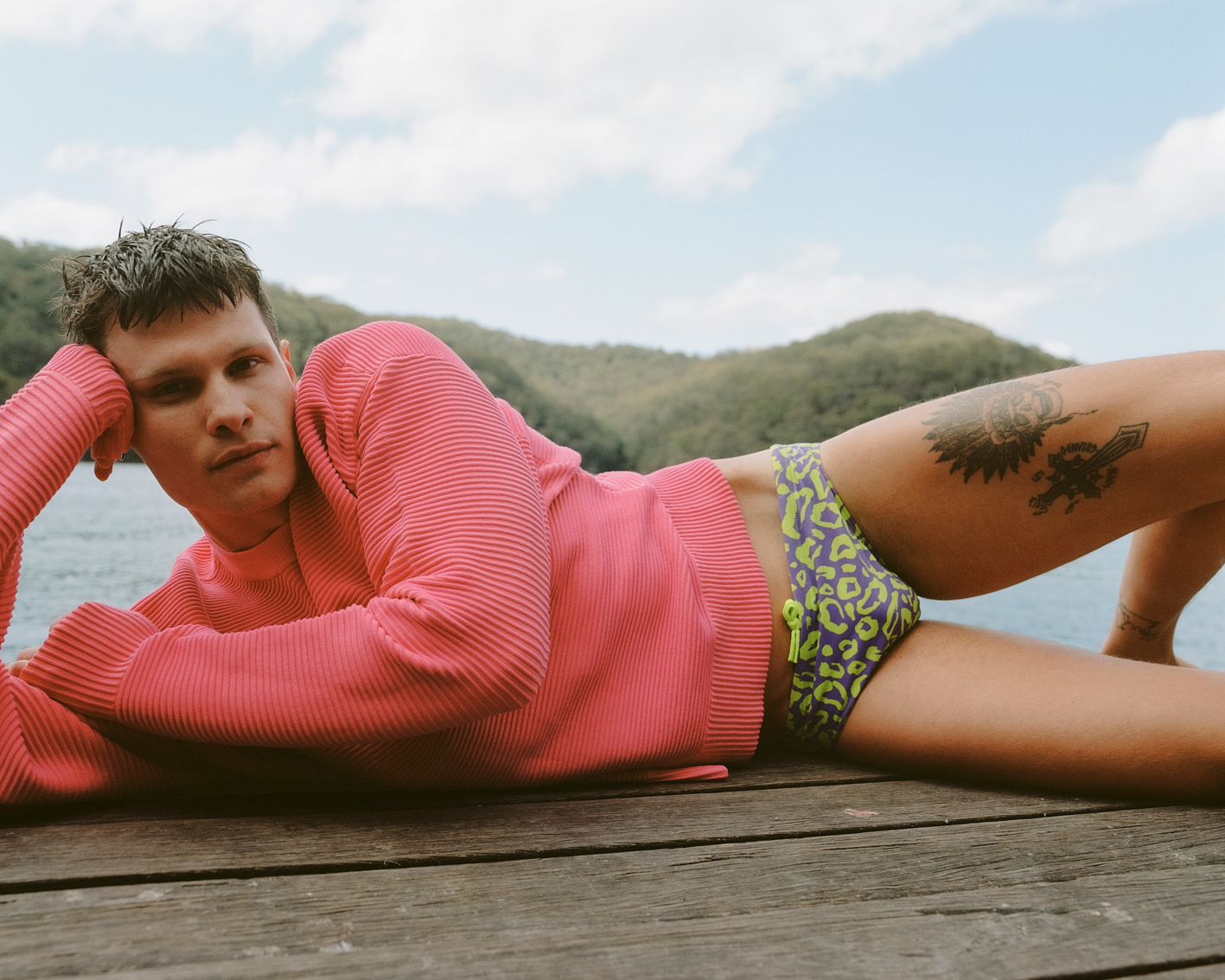 Young adult man in swimsuit and pink sweater relaxes on wooden dock with lake, mountain, and blue sky background during daytime.
