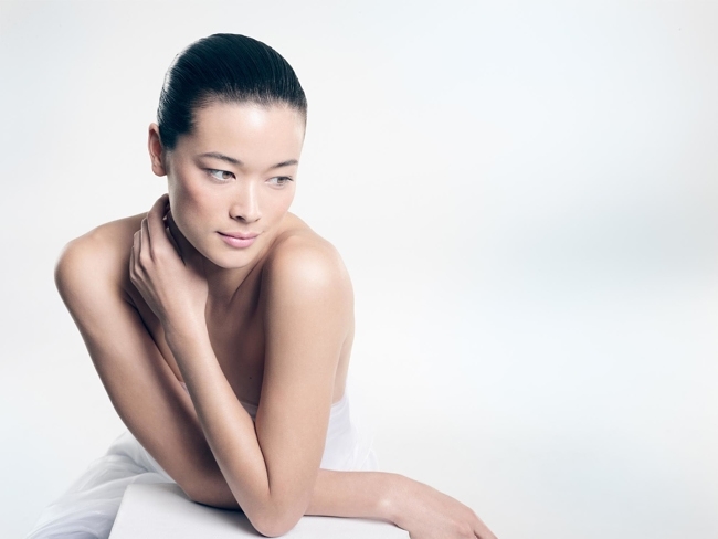 Asian woman portrait in bright studio with bare shoulders. Hands touching neck, glancing away. Clean minimalist aesthetic.