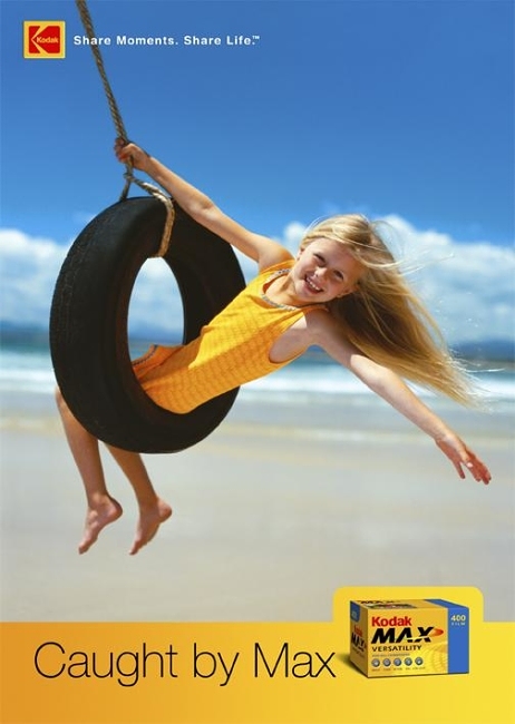 A young girl with long blond hair smiles joyfully as she swings on a tire swing suspended over a sunny beach with blue sky.