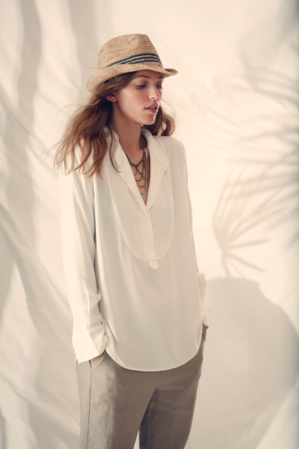 A young woman poses indoors, wearing a white blouse, straw hat, and grey pants, with soft lighting and a backdrop of palm tree shadows.