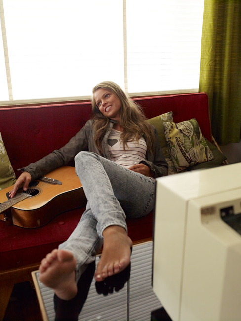 Woman relaxes on a red sofa with an acoustic guitar. She is barefoot with jeans and a jacket and looks up toward light from a window.