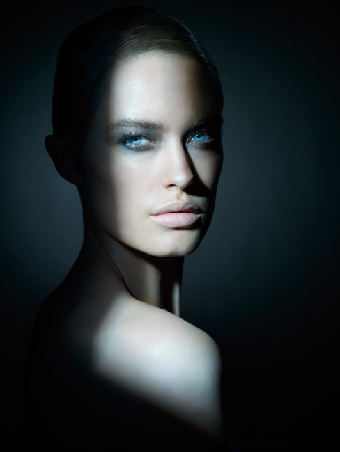 Headshot of a fair-skinned adult woman with blue eyes, partially lit by a vertical beam of light against a dark, shadowy background.
